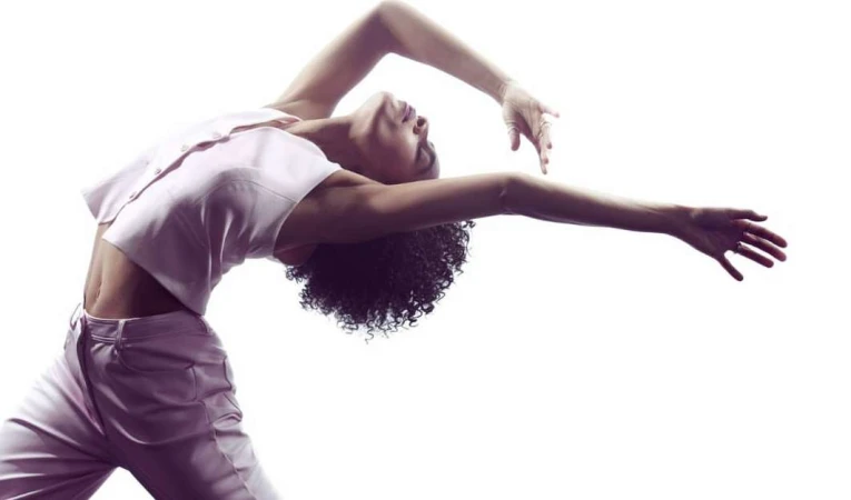 An athletic adult female does a modern dance pose wearing pink crop top and pants in a brightly lit minimalist photo studio.